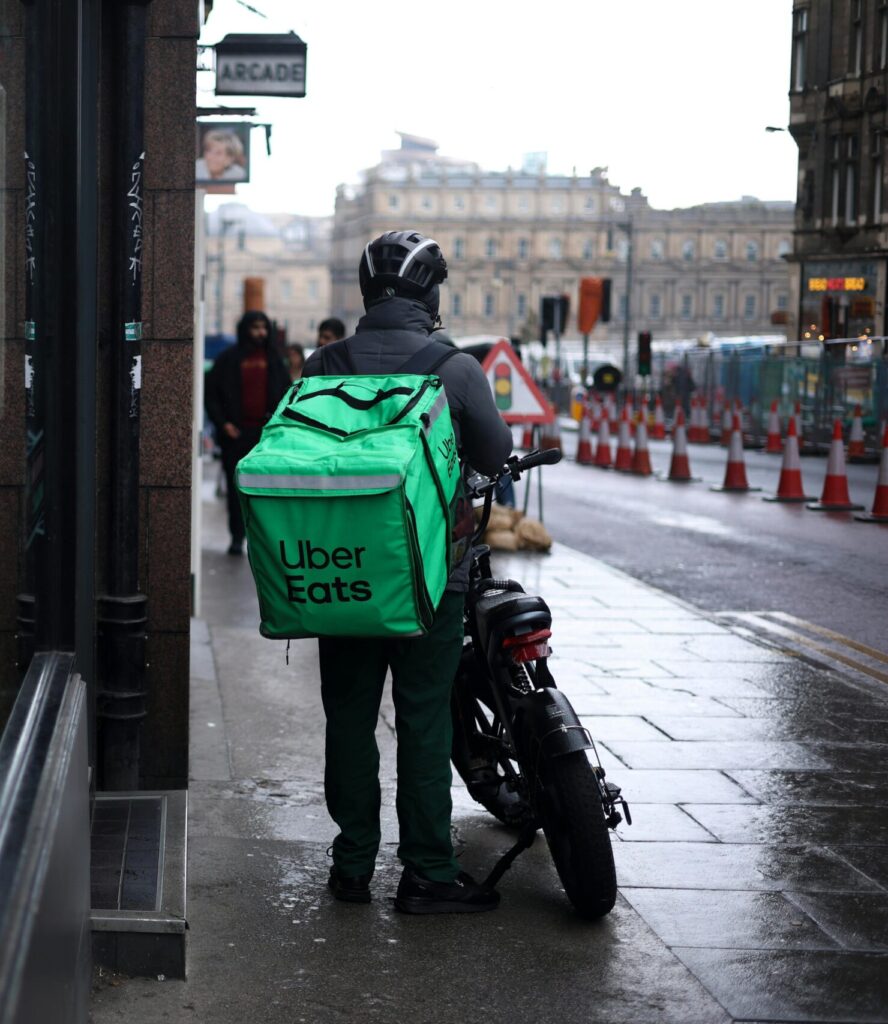 Uber Eats courier on motorbike in Edinburgh with mirror reflection on rainy street day.